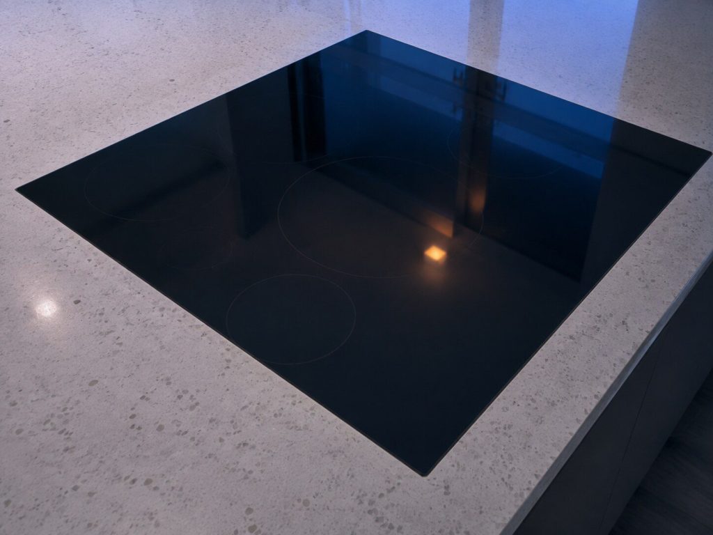 A black glass induction cooktop set flush into a quartz counter, photographed from above and slightly to the side so the ring outlines are only just visible in the reflection.