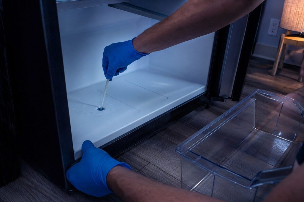A crisper drawer lifted out and set aside, a thin plastic blade held in gloved fingers clearing the small round drain hole at the back of the compartment floor, a film of water around it.