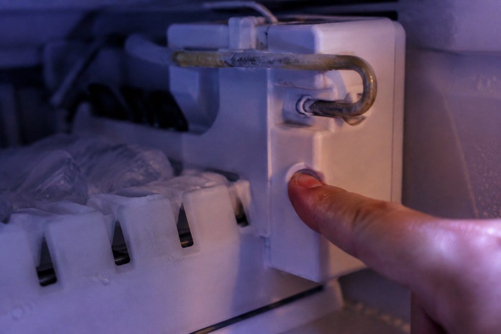 A finger pressed into the small recessed button on the side of an ice maker module inside a freezer compartment, the ejector arm above it.
