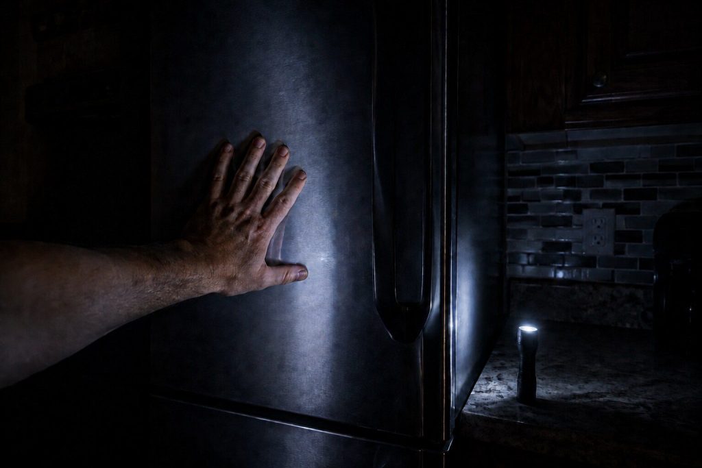 A hand pressed flat against a closed refrigerator door in a dark kitchen lit only by one flashlight standing on its end on the counter.
