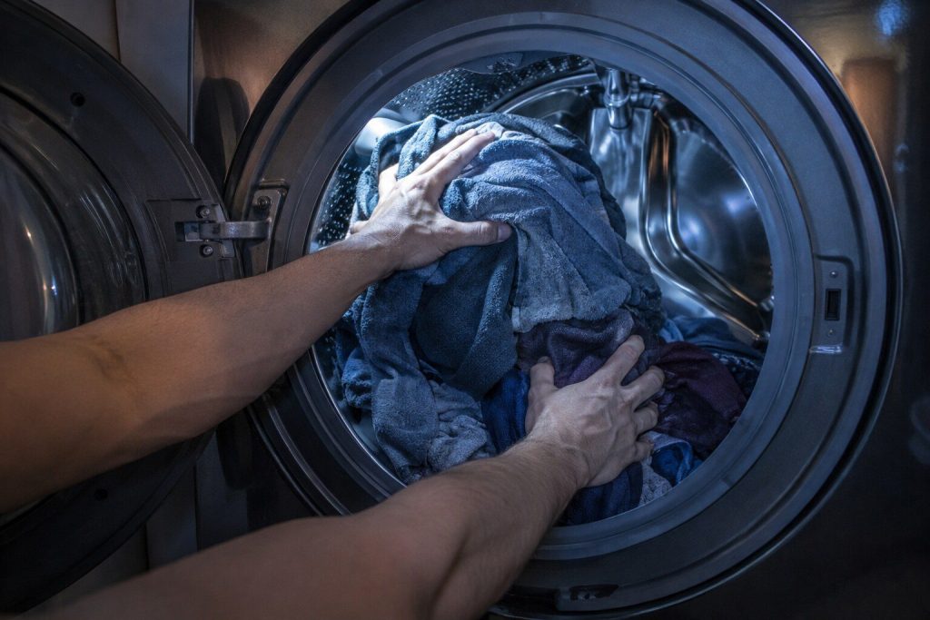 Two hands lifting a heap of wet laundry off one side of a washer drum and spreading it evenly around the wall of the drum.