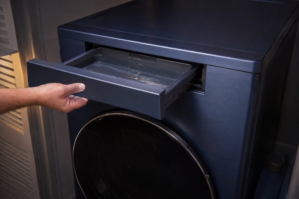 A tall brushed navy blue dryer with its condensate drawer drawn out at the top of the cabinet and held in one hand, water standing in it, the closed door below.