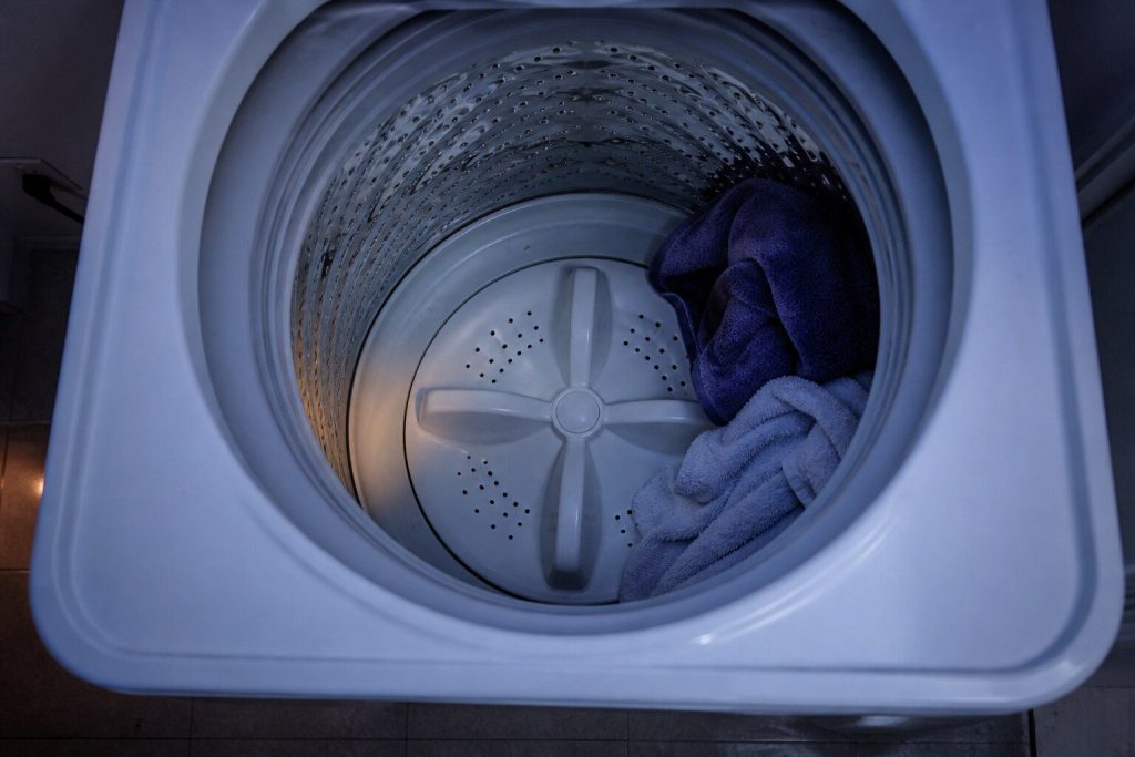Looking straight down into the open stainless tub of a white top load washer, the perforated wall running away below and the flat impeller plate centered at the bottom, a few towels against one side.