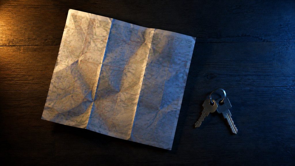 A folded paper map and a set of keys on a dark table at night, shot from above, one warm lamp to the side and cool blue fill.