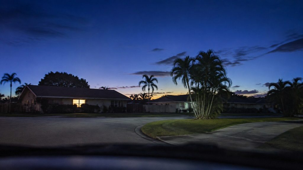 A South Florida residential street at dusk seen from a parked vehicle's point of view — low houses, palms, a deep blue sky with the last warm light behind them, one lit window.