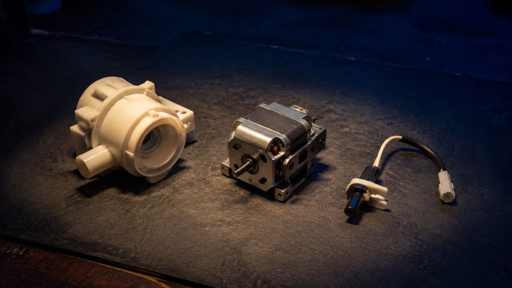Three small unbranded appliance components laid out on a clean workbench mat under a single work light — a pump housing, a small motor and a sensor with a lead.