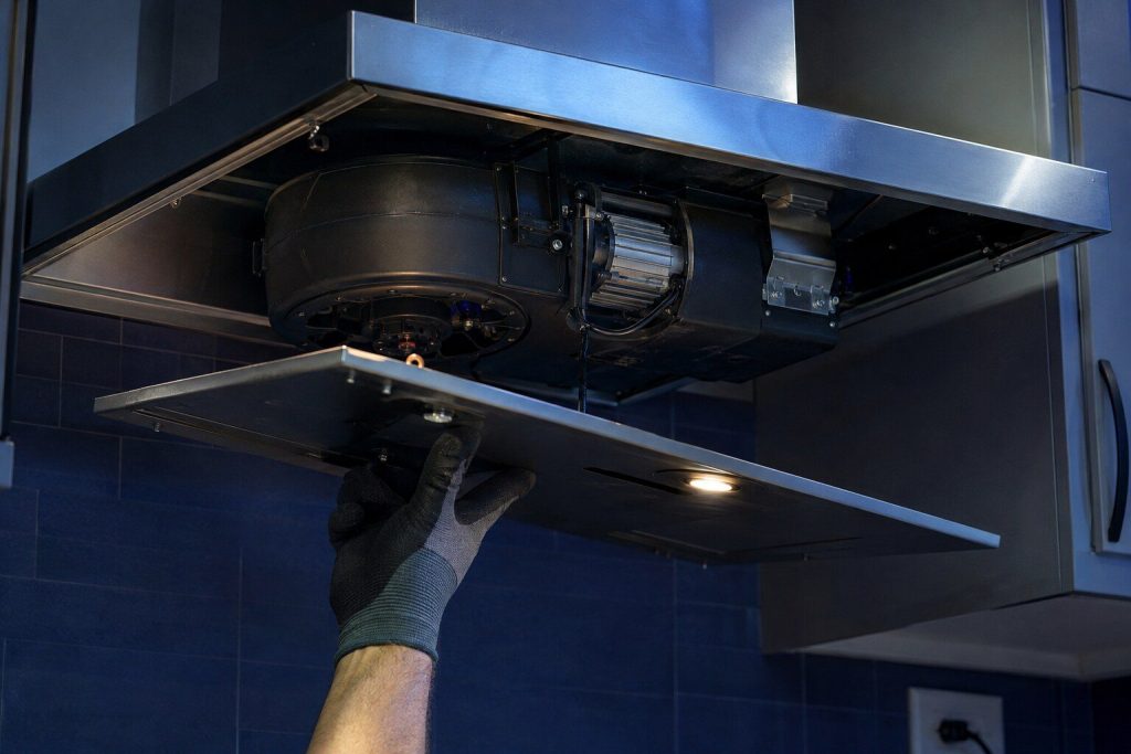 The underside of a wall hood with its lower cover and baffle filters taken out, the blower housing and its motor exposed inside the canopy, a gloved hand supporting the cover.
