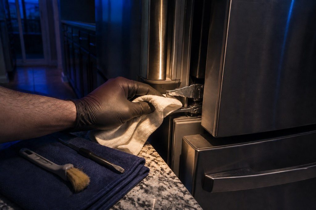A gloved hand drawing a clean cloth along the hinge and door channel of a stainless appliance, a soft brush and a small screwdriver lying on a folded mat beside it.