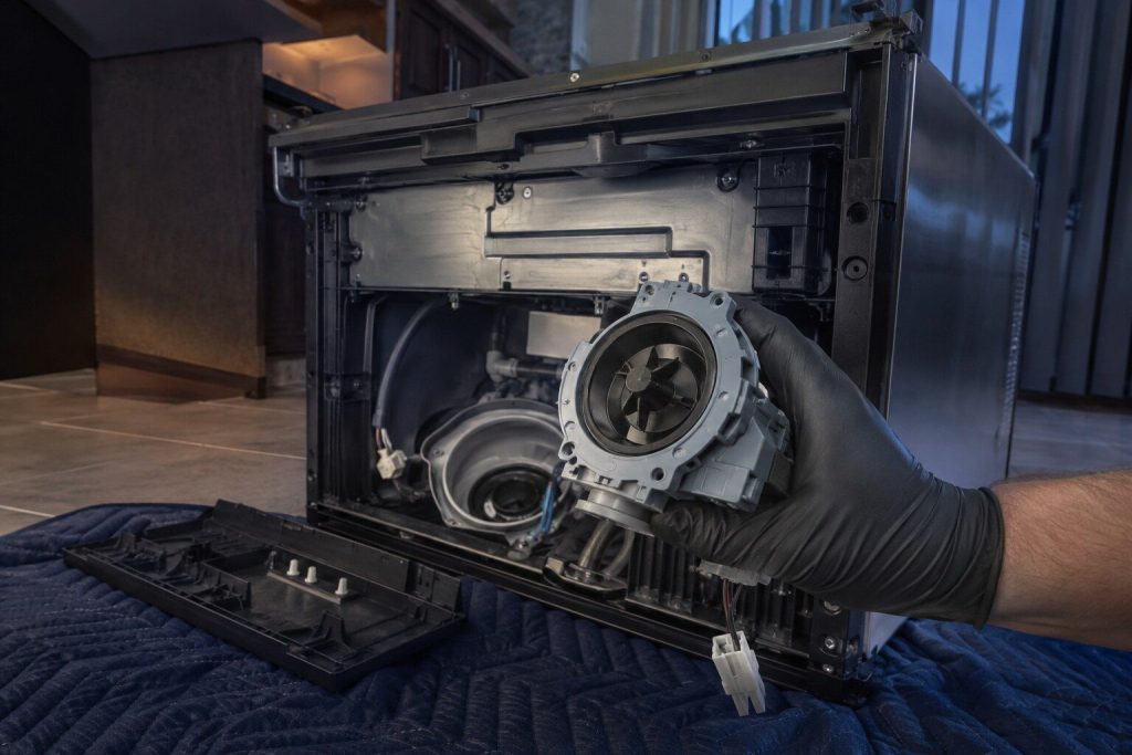 A dishwasher laid on its back on a blanket with its base cover off and the drain pump lifted clear of the sump on a gloved hand, the impeller housing turned to the camera.