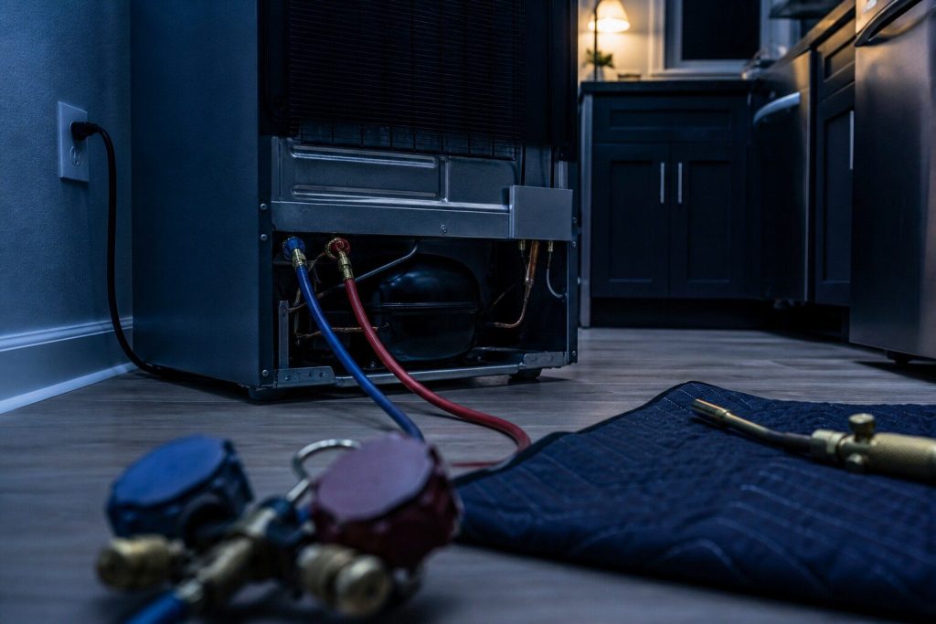 Two charging hoses connected to the process ports on the back of a refrigerator, the gauge bodies lying out of focus in the foreground with their faces turned away, a brazing torch and a heat mat on the floor beside them.
