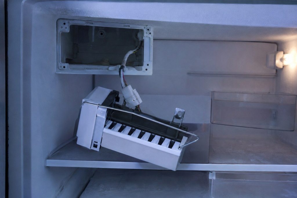 An ice maker module unclipped from the freezer wall and hanging on its own wiring harness, its ejector arm and mounting bracket clear, the empty bay behind it.