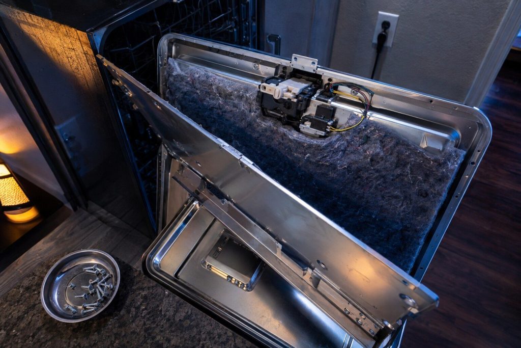 The inner panel of a dishwasher door unscrewed and lifted away with the insulation folded back, the latch assembly and its switch exposed at the top edge and the screws in a magnetic tray.