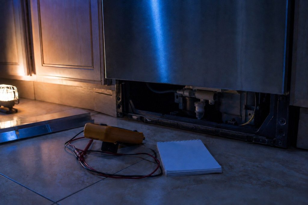 A digital multimeter and a small notepad set down on a kitchen floor beside an appliance whose lower access panel is off, the meter's own face turned away from the camera.