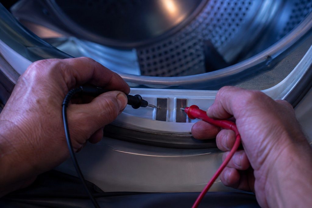 Two meter probes held against the pair of metal sensor strips just inside the front of a dryer drum, the leads running out of frame.