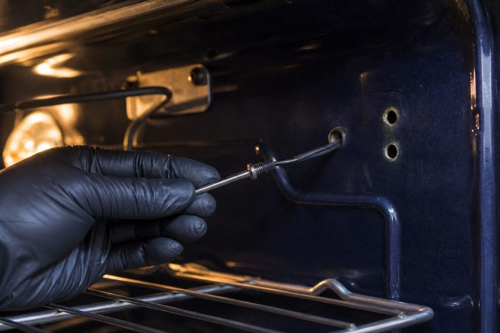 The slim oven temperature probe unscrewed from the cavity wall and pulled clear on its lead, held in gloved fingers with the two empty screw holes behind it.
