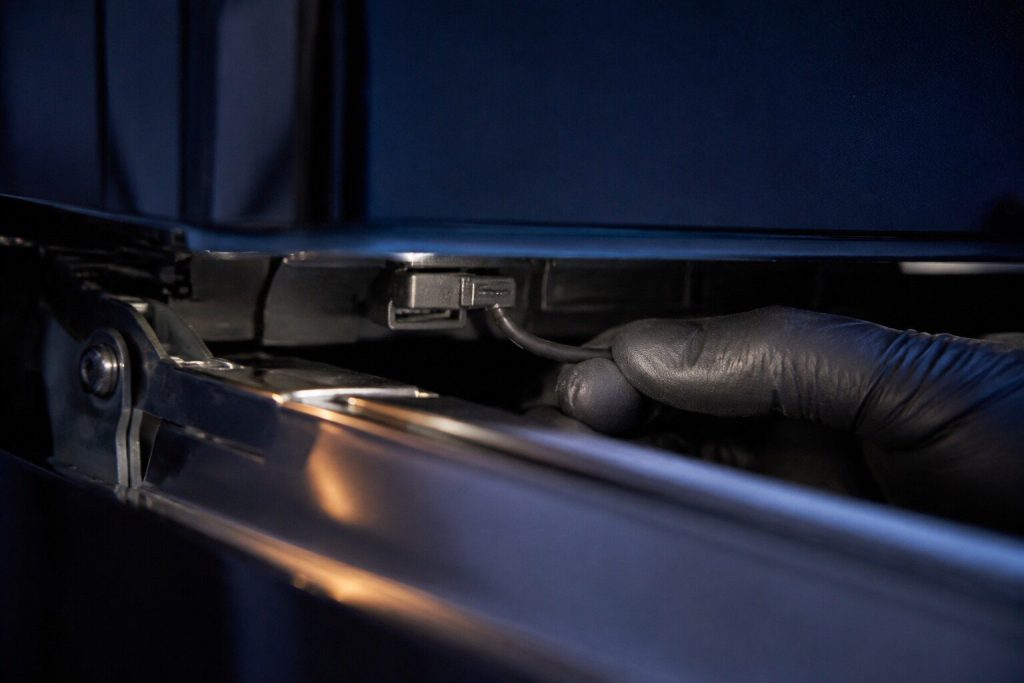 A short service cable plugged into a small port on the underside of an appliance control panel with the fascia trim swung down, gloved fingers steadying the connector.