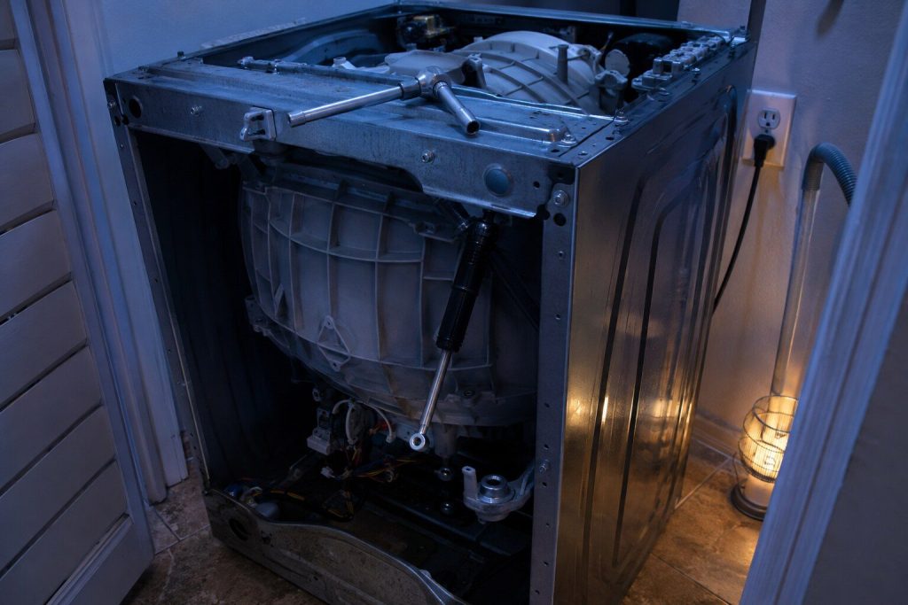 A washer with its top and front off, one suspension damper rod unbolted at the lower mount and swung clear of the outer tub, a socket wrench resting across the frame.