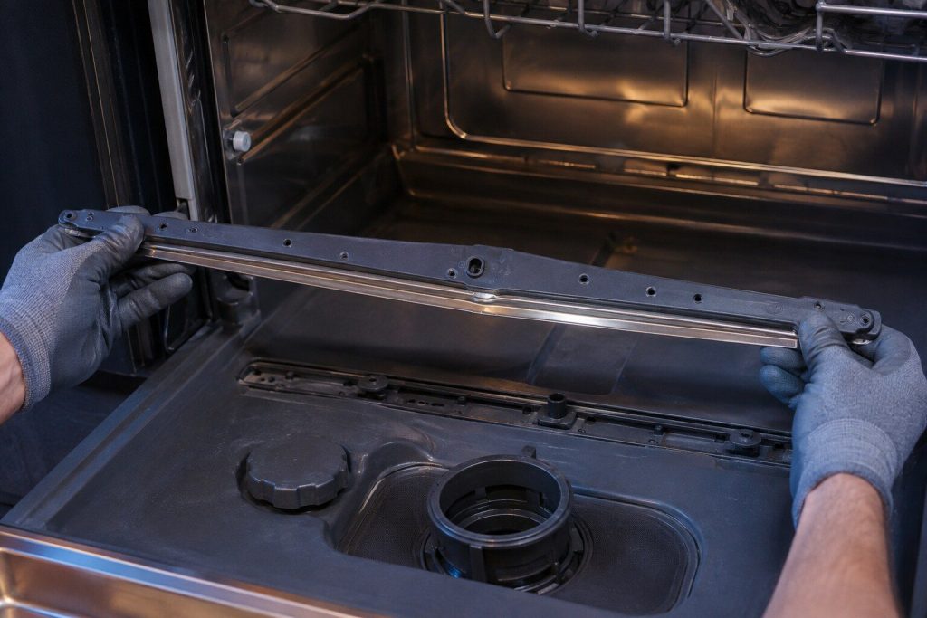A long flat wash bar and its reflector strip lifted up out of the floor of a dishwasher tub on two gloved hands, the empty track it sits in visible below.