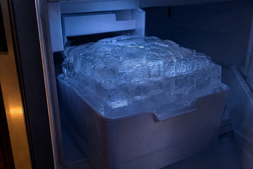 An ice maker compartment where the loose cubes have frozen into one solid clear mass filling the whole bucket and standing proud of its rim.