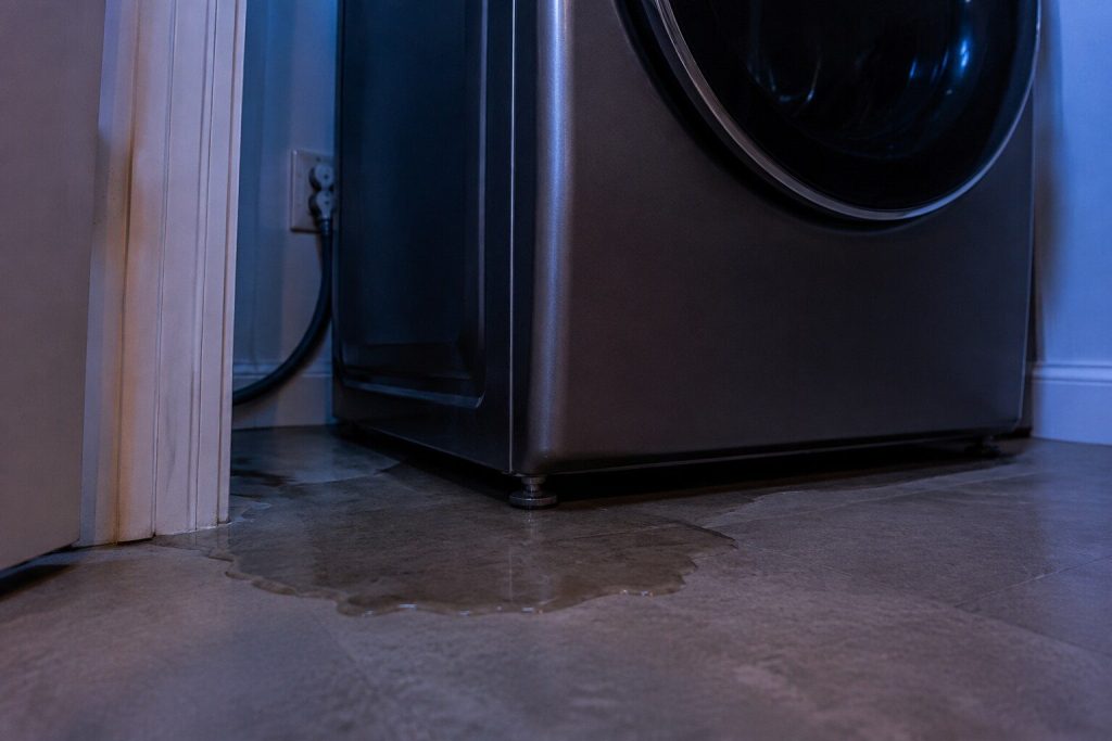 A dark wet patch spreading across a laundry closet floor from beneath the front left corner of a washer, the leveling foot standing in it.
