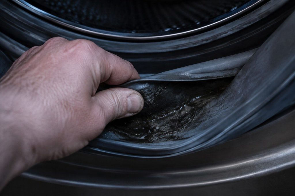A hand folding back the rubber boot gasket of a front load washer to show the dark slimy residue lying in the fold.