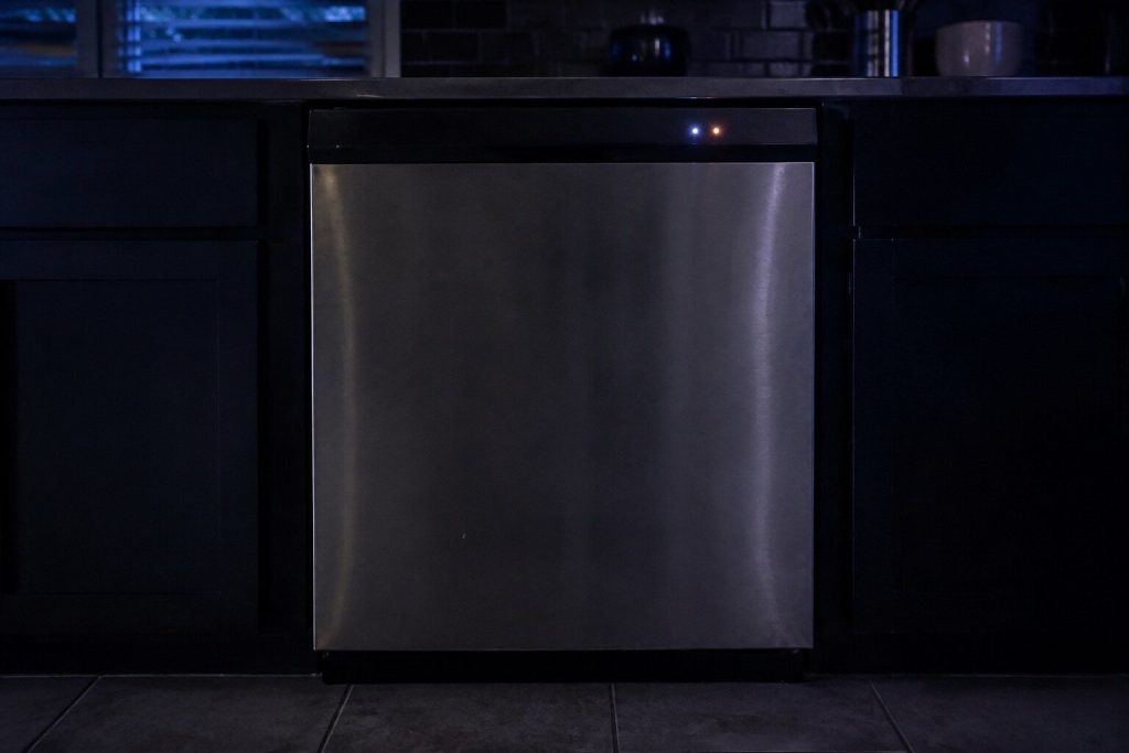 A dishwasher front seen straight on in a dark kitchen with two small round indicator lamps lit on its top edge and everything else black.